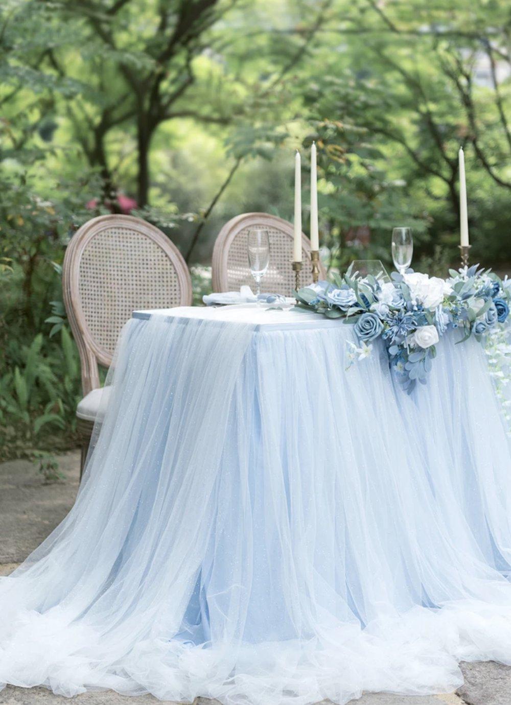 Outdoor table surrounded by greenery with a sky blue tulle tablecloth, French style chairs, blue and white floral runners and white candles with glassware