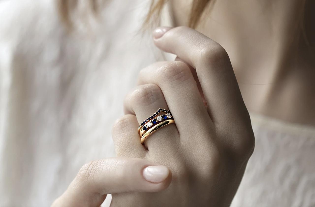 Close-up of white woman's hand with a nude manicure wearing a gold band, sapphire and diamond band and a wishbone shaped sapphire studded gold ring