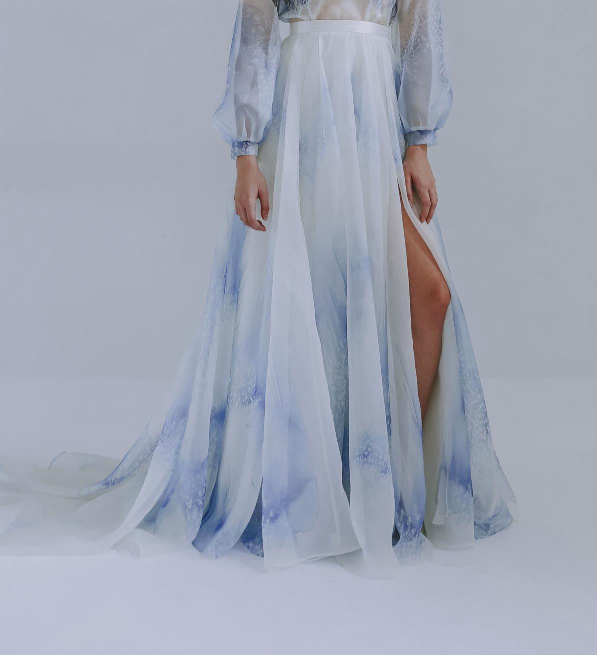 White woman wearing a flowing blue and white bridal skirt with a split and a sheer long sleeved blue and white blouse tucked in at the waistband