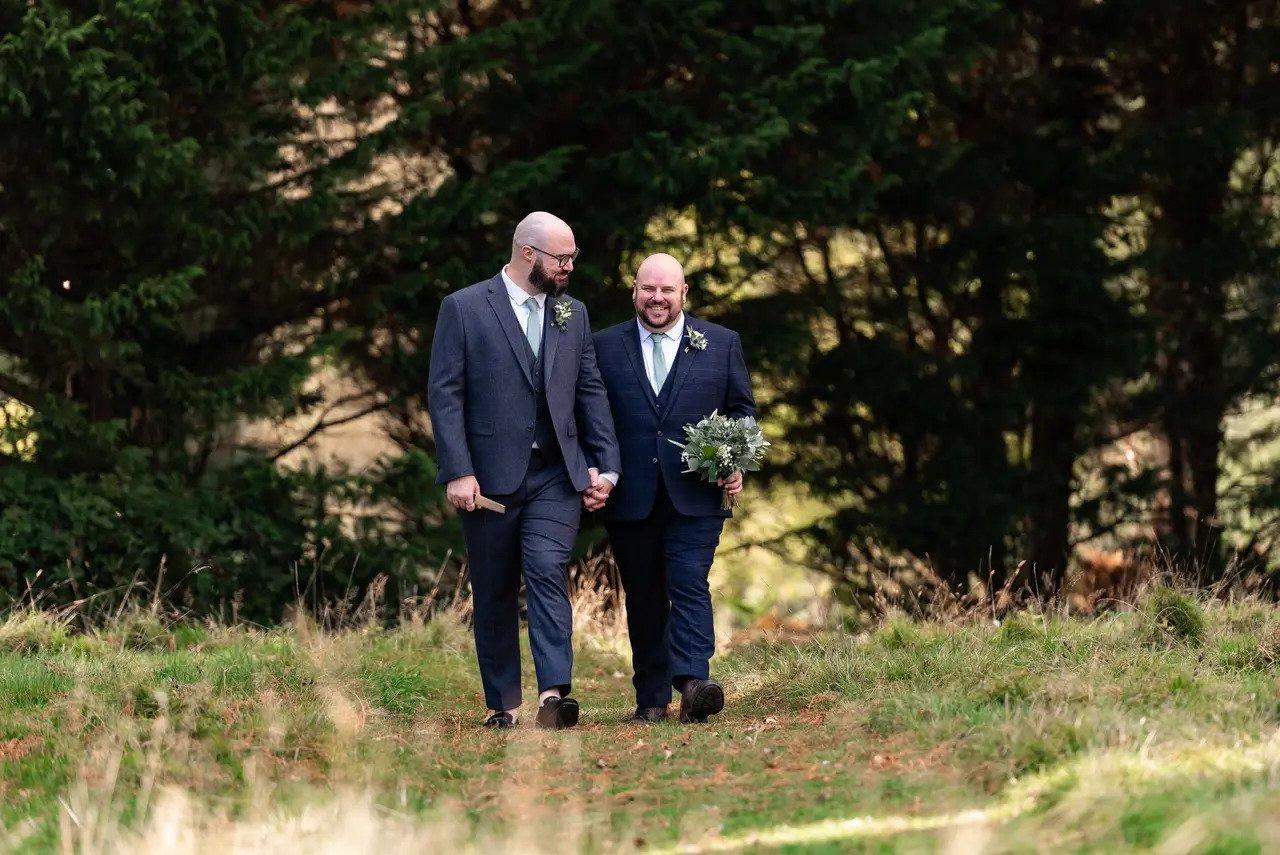 Two grooms walking outside in the grounds of a wedding venue, hand in hand. One of the grooms carries a bouquet of flowers