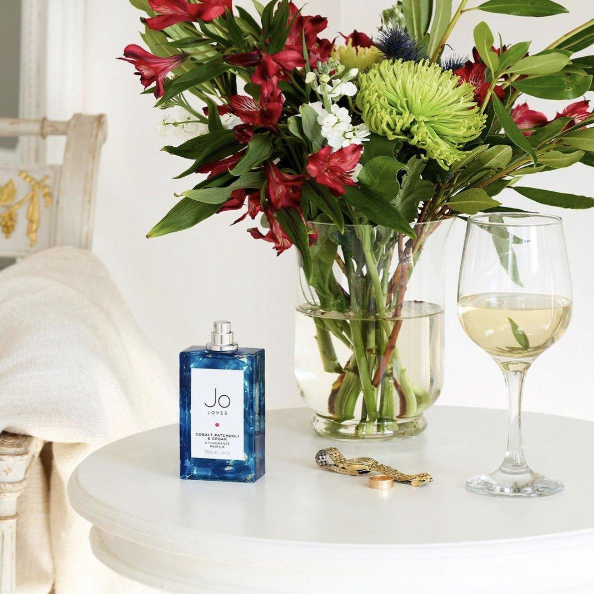 Blue rectangular Jo Loves Cobalt Patchouli & Cedar fragrance bottle on a white bedside table next to a vase of red and white flowers, a gold watch and a glass of white wine