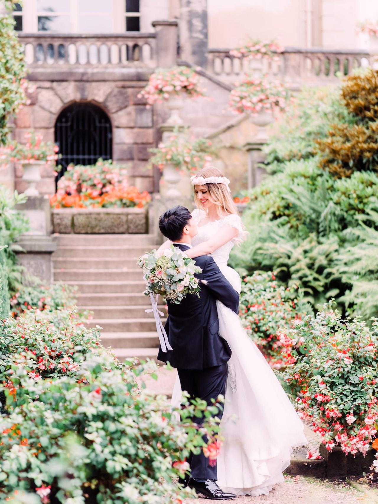 Groom lifting bride up in front of a villa style venue with garden full of greenery