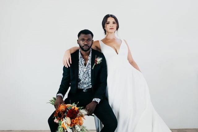 Groom sitting in a chair holding a bouquet with a bride standing next to him with her arm around her shoulders, both looking seriously at the camera