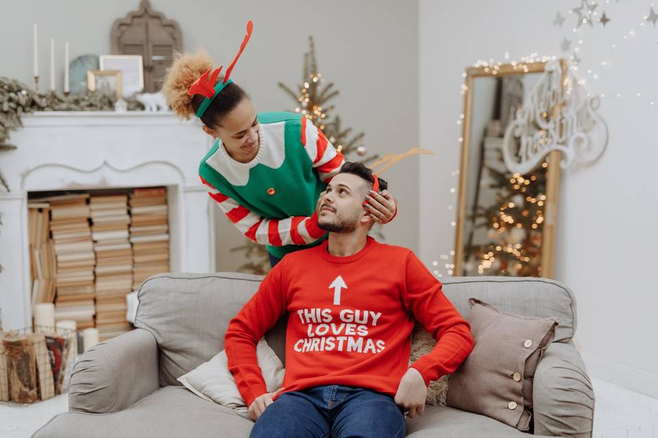 Man in a Christmas jumper reading 'this guy loves christmas' sitting on a sofa whilst a woman dressed as an elf is behind him tenderly touching his head