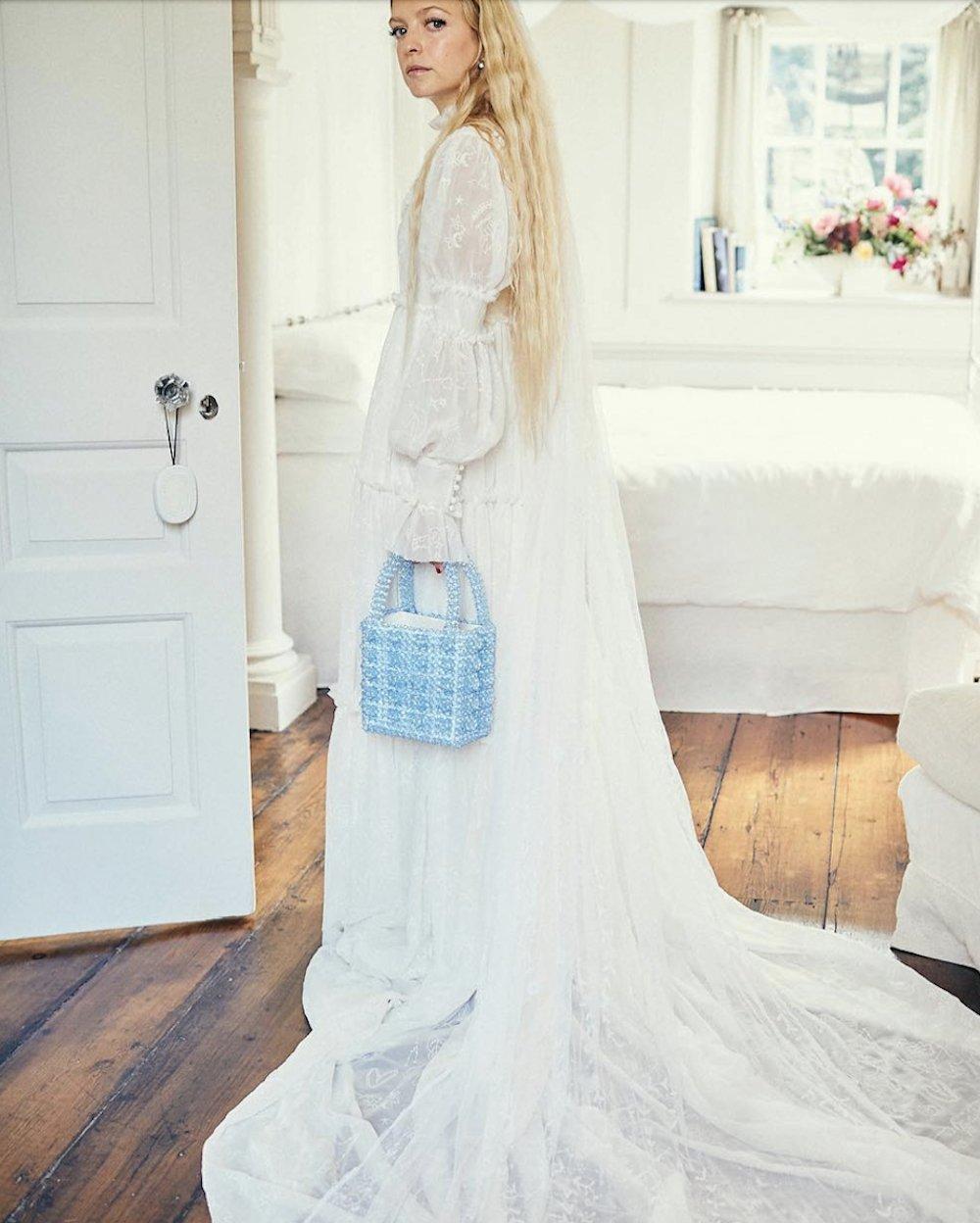 White woman with flowing wavy blonde hair in a white bathroom wearing a white high necked Victorian style gown earring a light blue beaded bag in her left hand