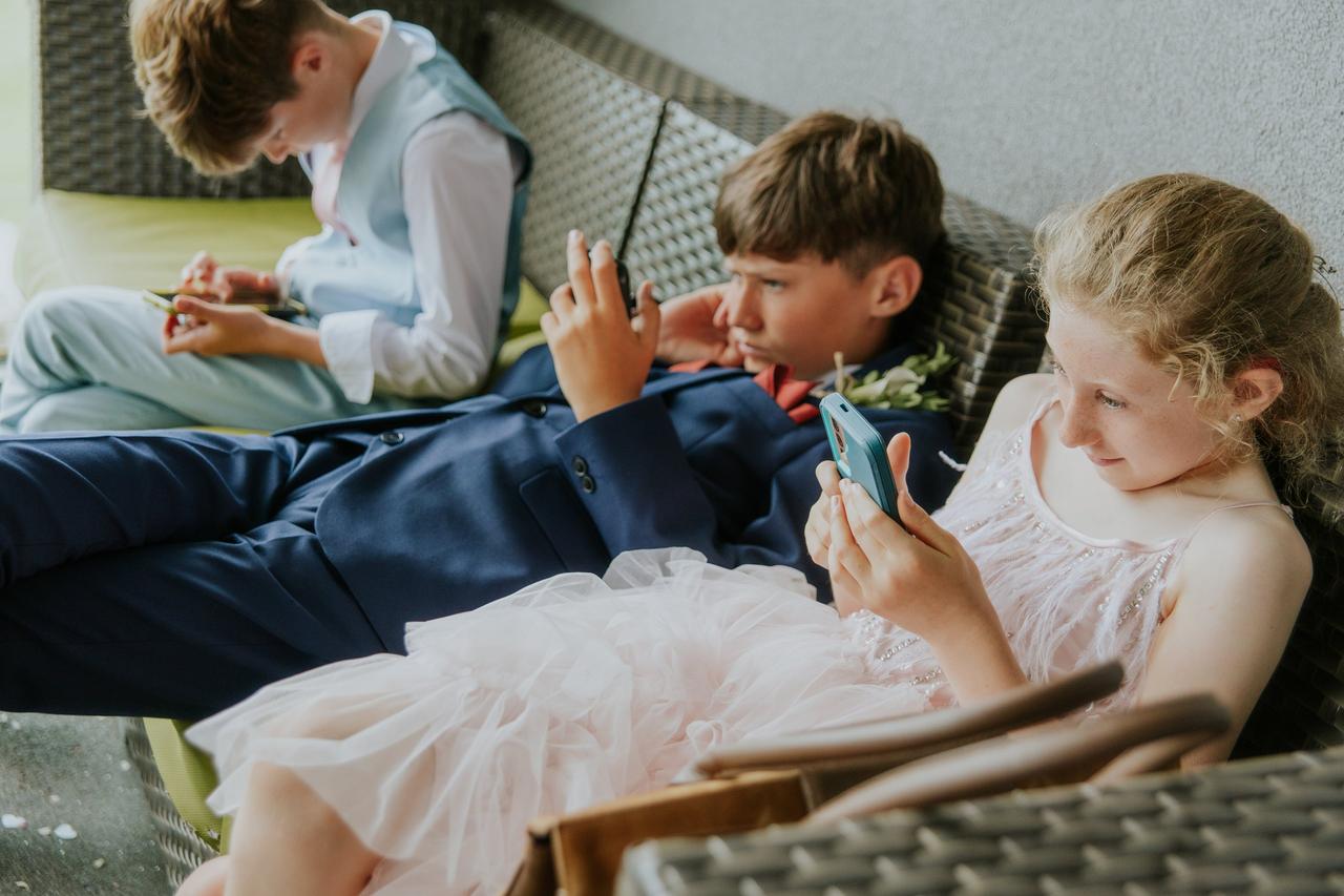 Three children in wedding attire leaning back on chairs looking bored as they read and play games on their phones.