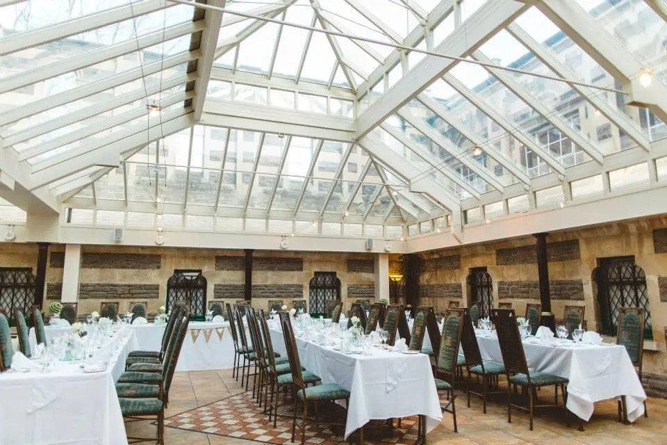 Room with glass roof set for wedding  breakfast with partially brick walls, long white tables and green chairs