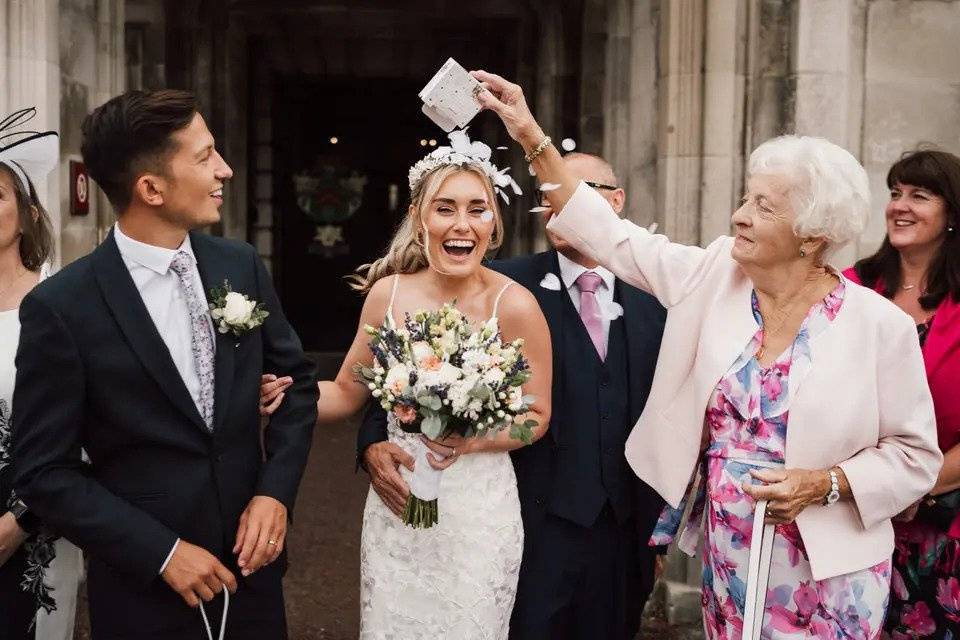 Bride and groom laughing as an elderly lady tips confetti directly on to the bride's head from a packet