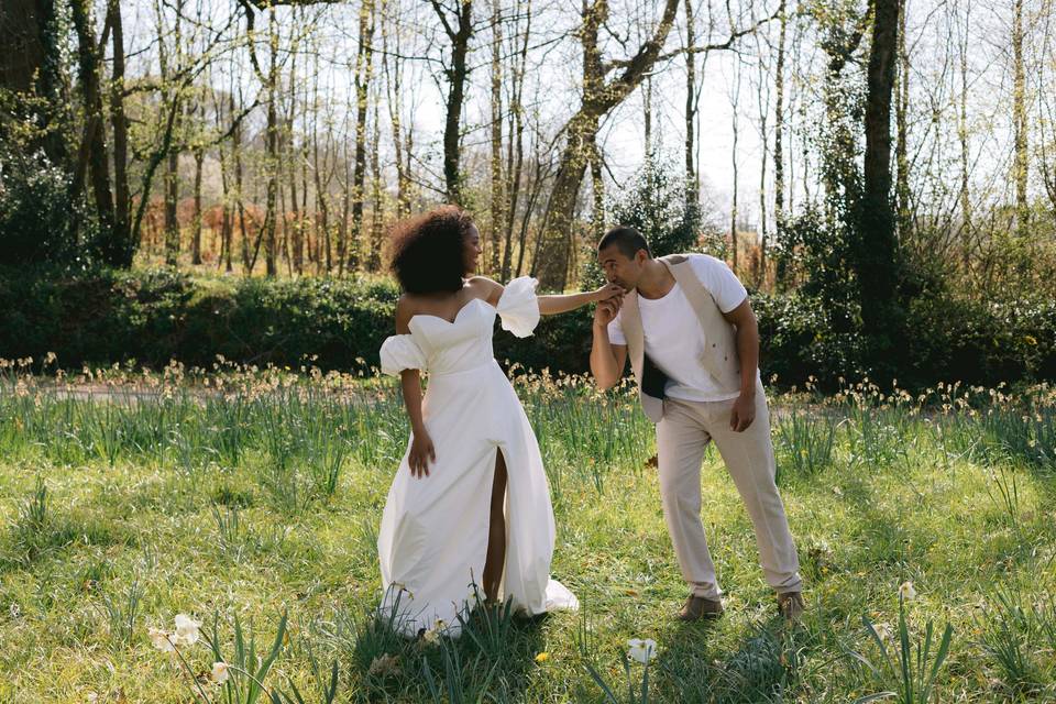 A groom standing in a meadow of flowers kissing the hand of a bride in a white dress