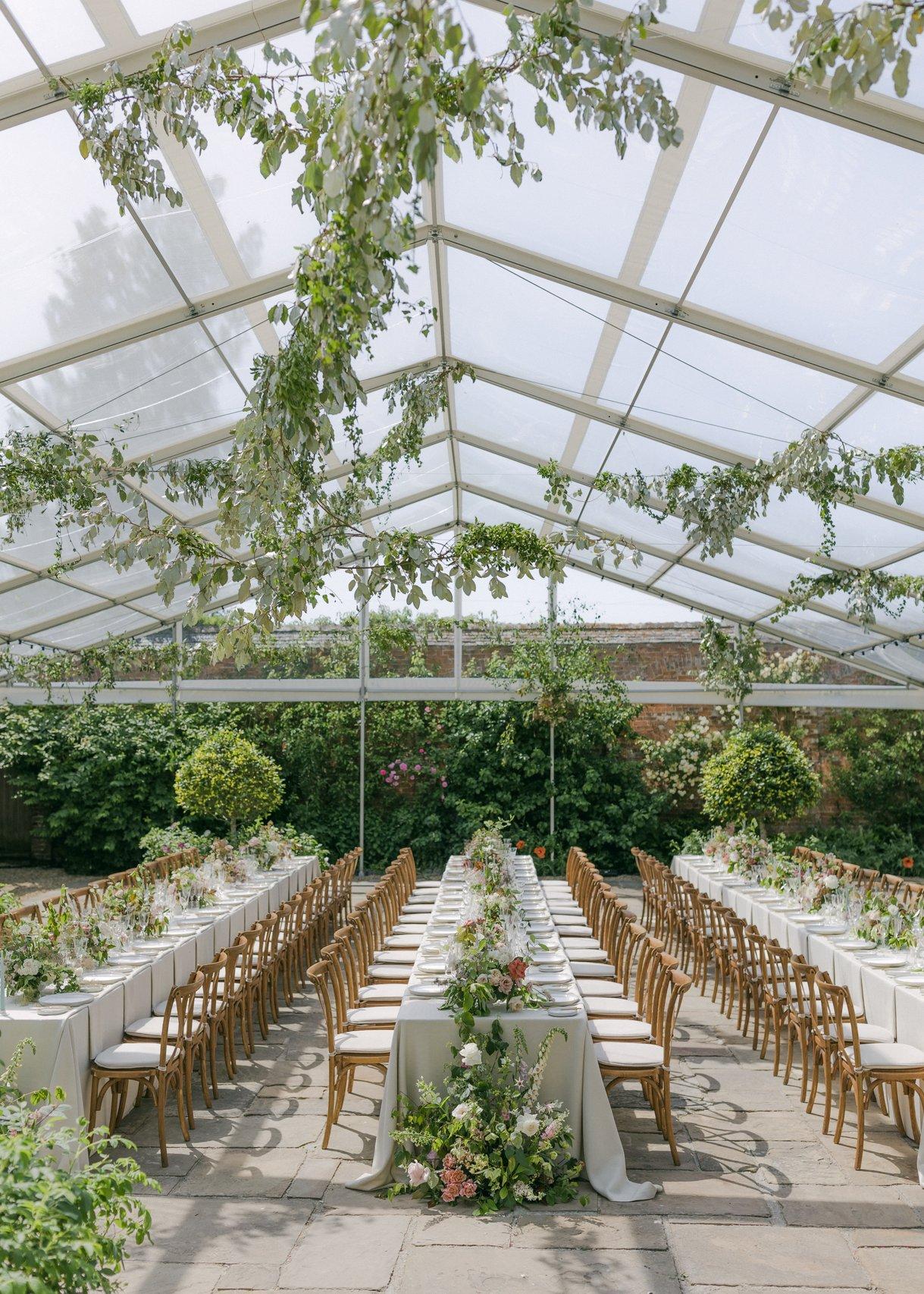 A glass conservatory wedding venue set up for a wedding dinner with long tables and lots of foliage and flowers.