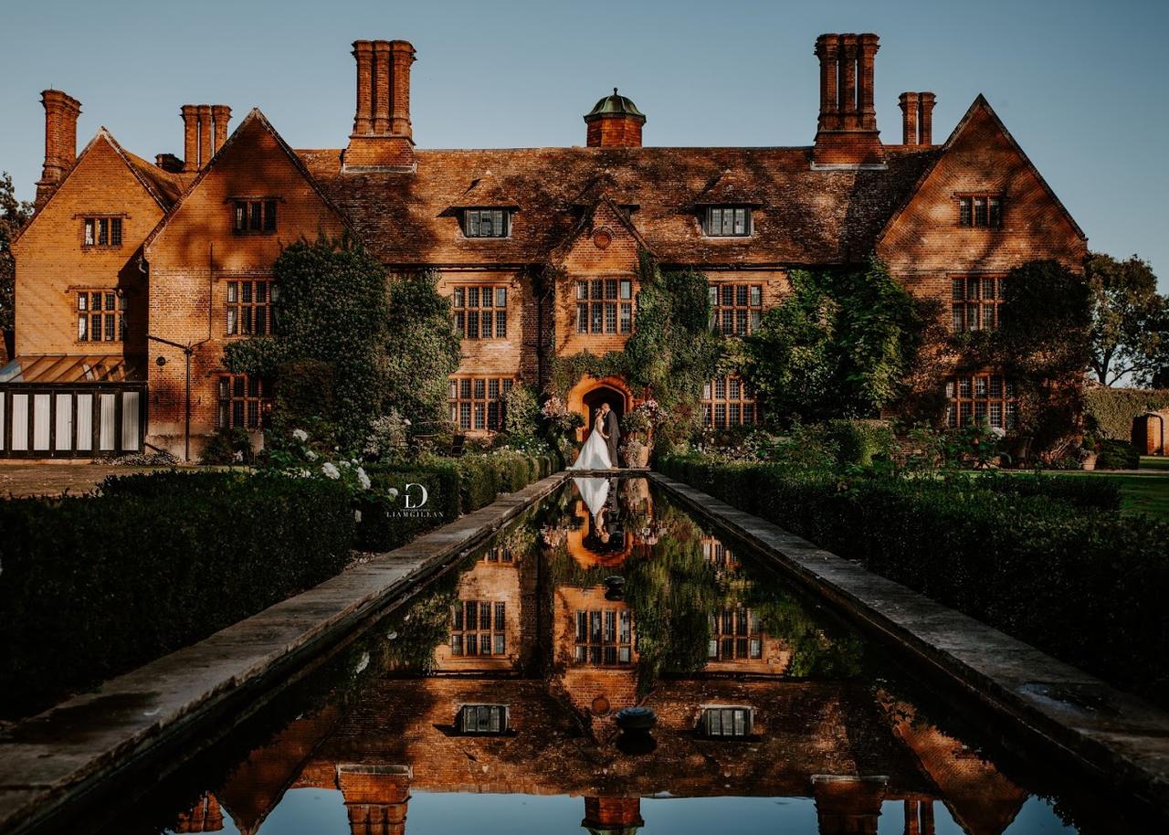 Exterior of an elizabethan style manor house with a pond in front and a bride and groom