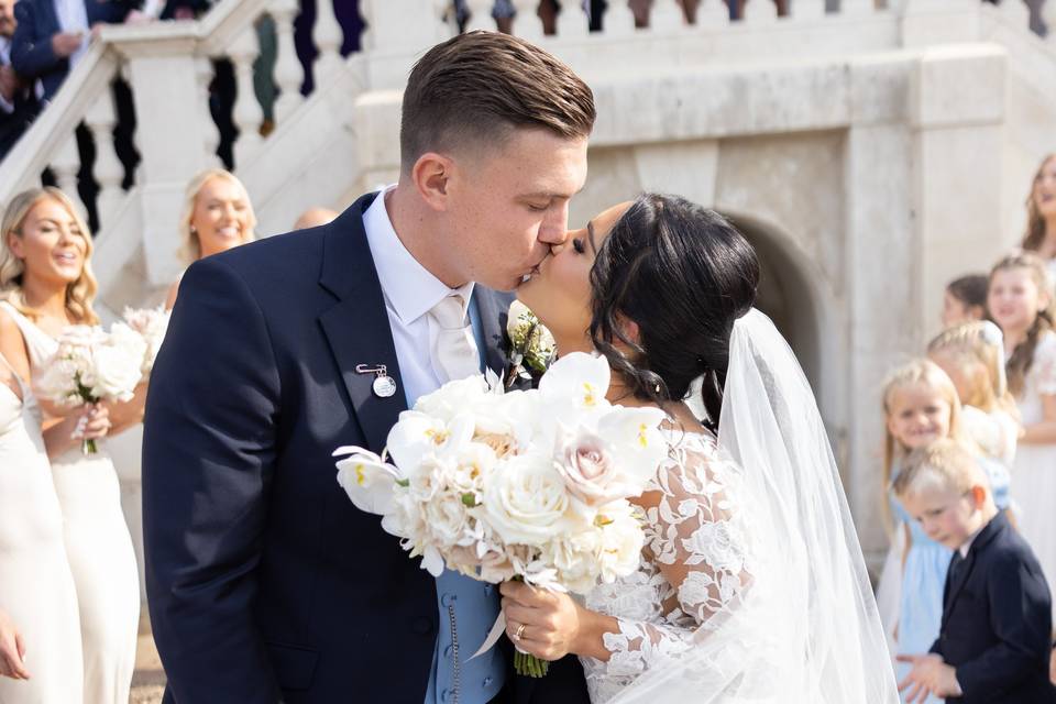 a groom in a navy suit kisses his bride as she holds her bouquet outside botleys mansion