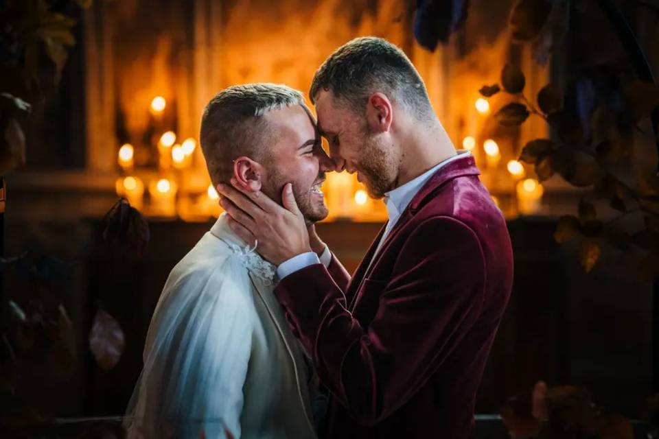 Two grooms kissing in a candle-lit room - one wearing a red velvet suit and the other in a cream blazer