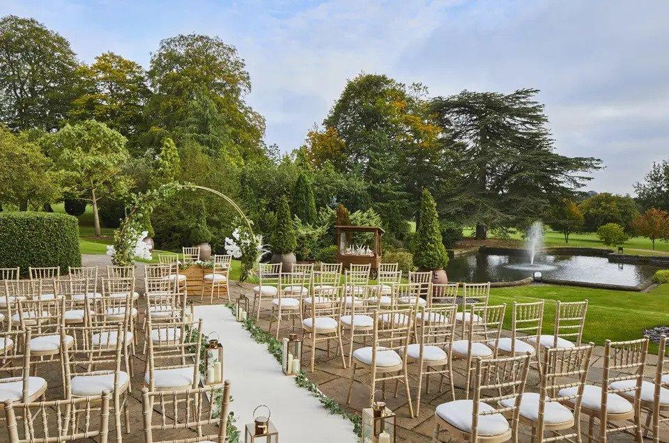 Outdoor wedding set up with wooden chairs, pillar candles, foliage decor, and a floral hoop surrounded by trees beside a fountain