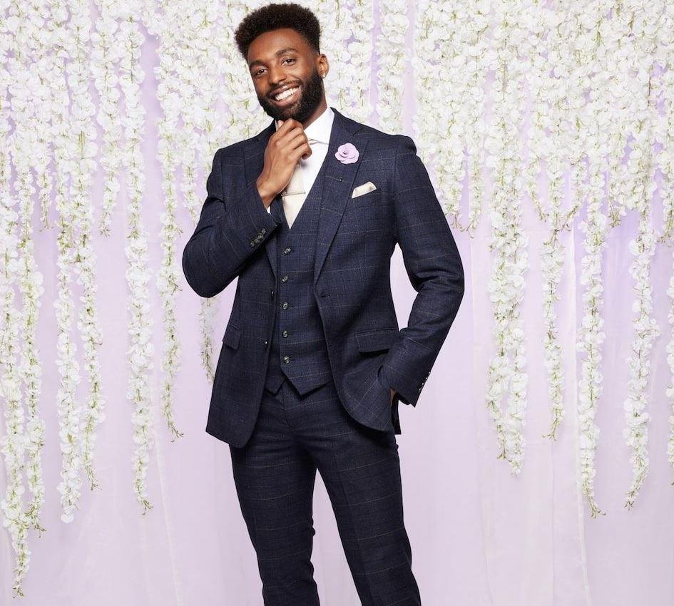 mafs uk 2023 contestant paul wears a smart dark blue checkered suit and smiles in front of a lilac backdrop with white hanging flowers