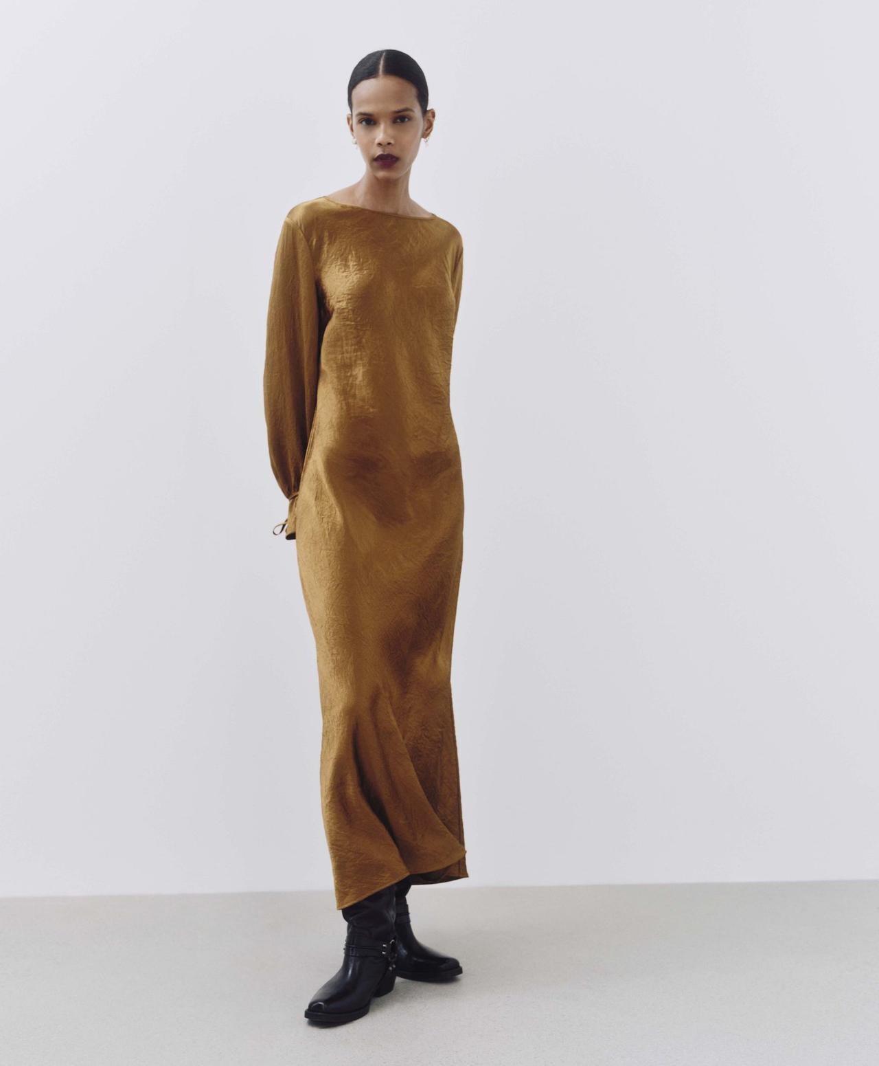 A model wearing a gold satin midi dress with long sleeves and flared cuffs. She is wearing it with chunky black boots for a more relaxed look.
