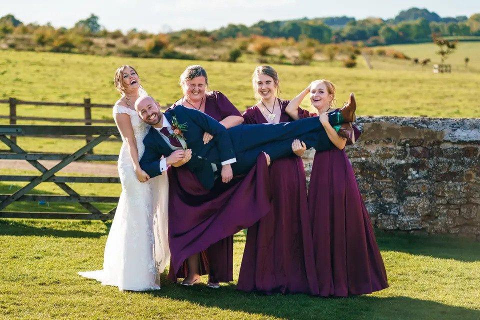 bride and bridesmaids lift up groom in a countryside field