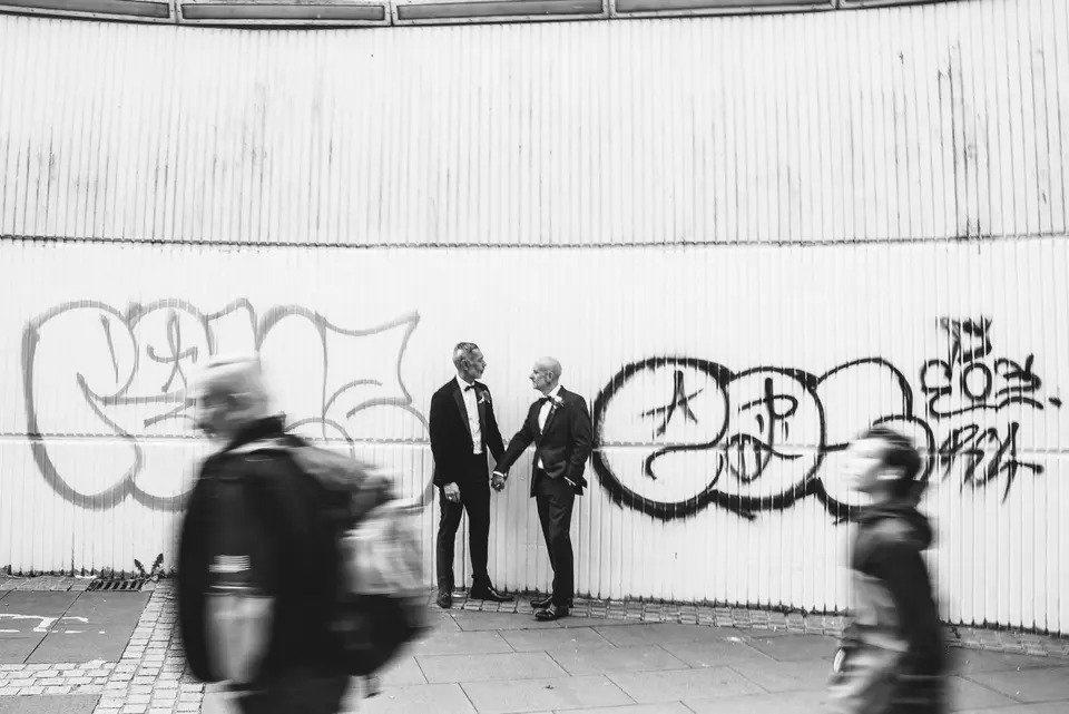 Black and white photo of two grooms holding hands in front of a wall covered in graffiti
