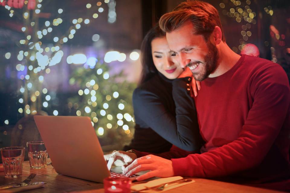 Couple smiling as they lean over a laptop in front of twinkling lights