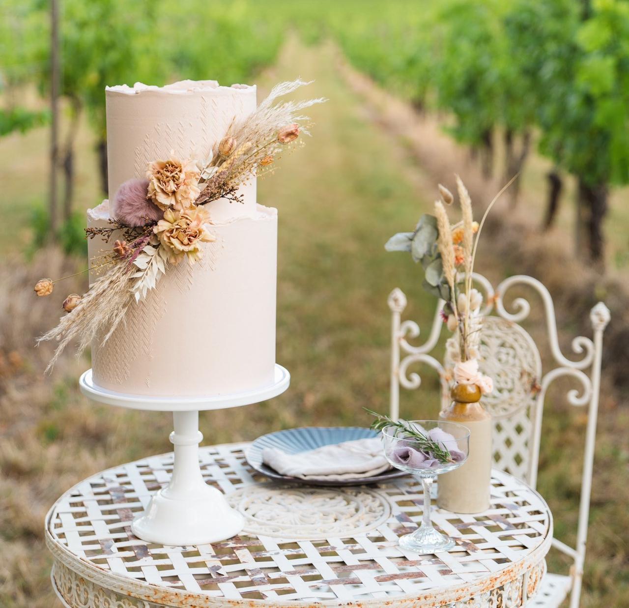 A simple two tier wedding cake decorated with a spray of dried flowers on a table at a vineyard wedding venue.