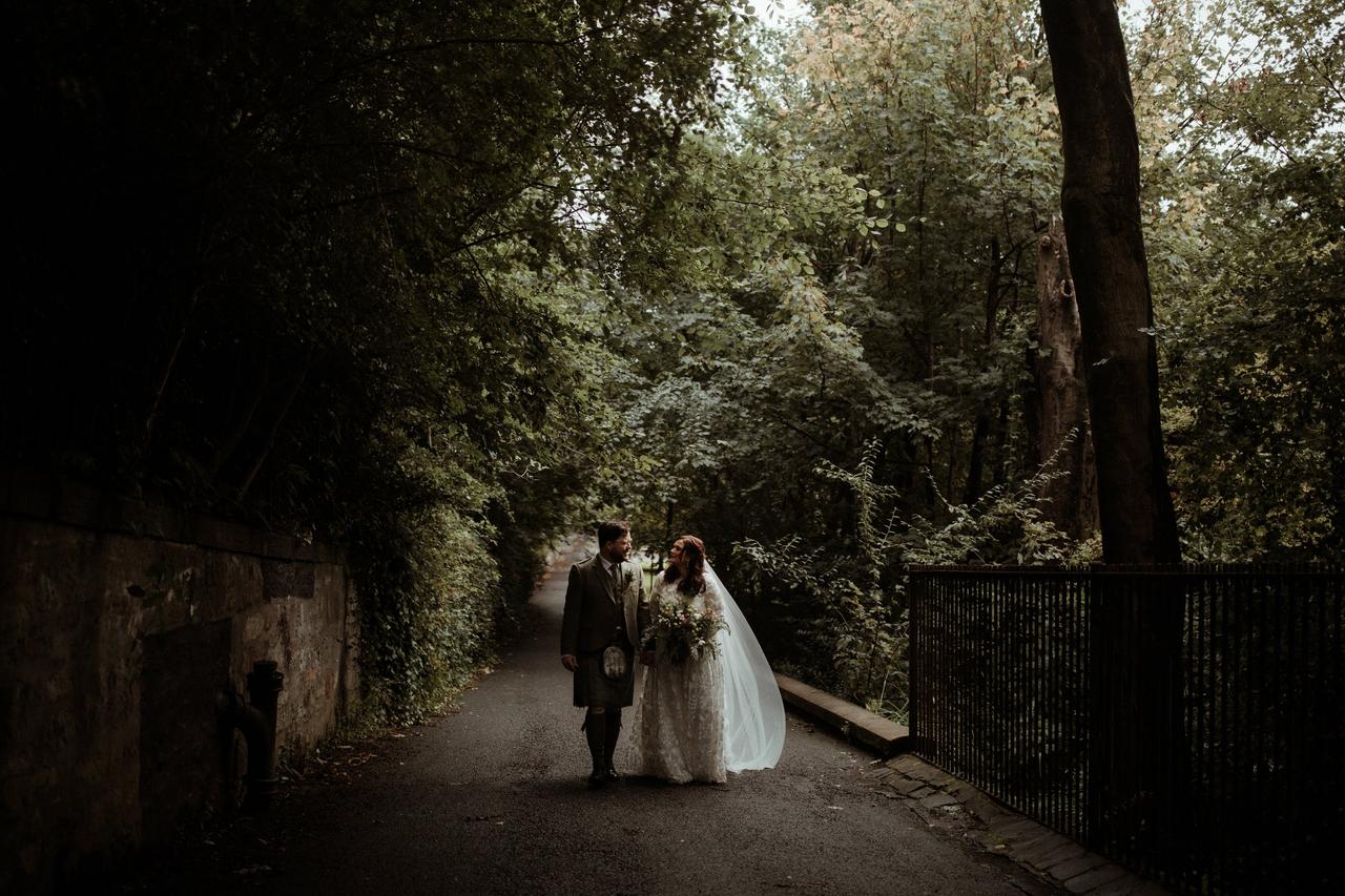 White newlyweds walking down a park path surrounded by green leaves with the groom wearing a green tweed suit and a kilt and the bride wearing a floral embroidered long-sleeved dress and white veil