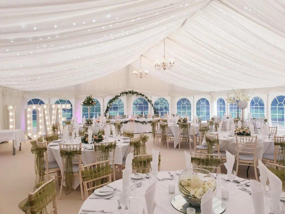 Marquee dressed for wedding meal with white round tables, wooden chairs with green sashes, chandeliers, illuminated letters spelling LOVE and a floral arch