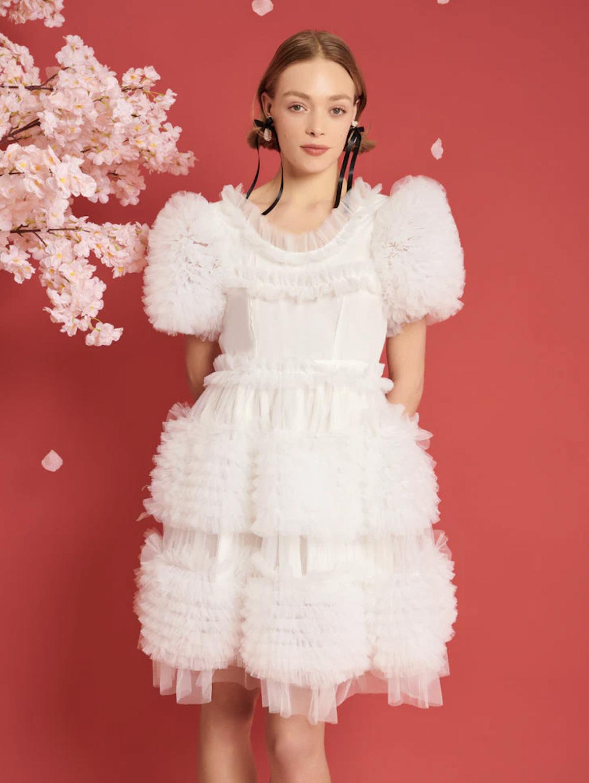 woman standing in front of floral background wearing a short ruffled wedding dress with black ribbons in her hair