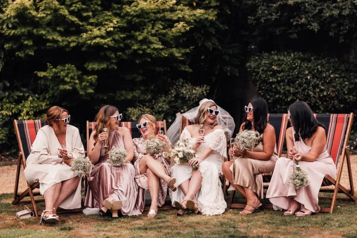A bride and her bridesmaids sitting on deck chairs with sunglasses on drinking and laughing