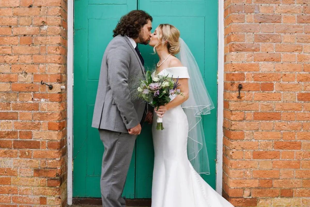a bride and groom in a wedding dress and suit kissing in front of a green door and light brick wall