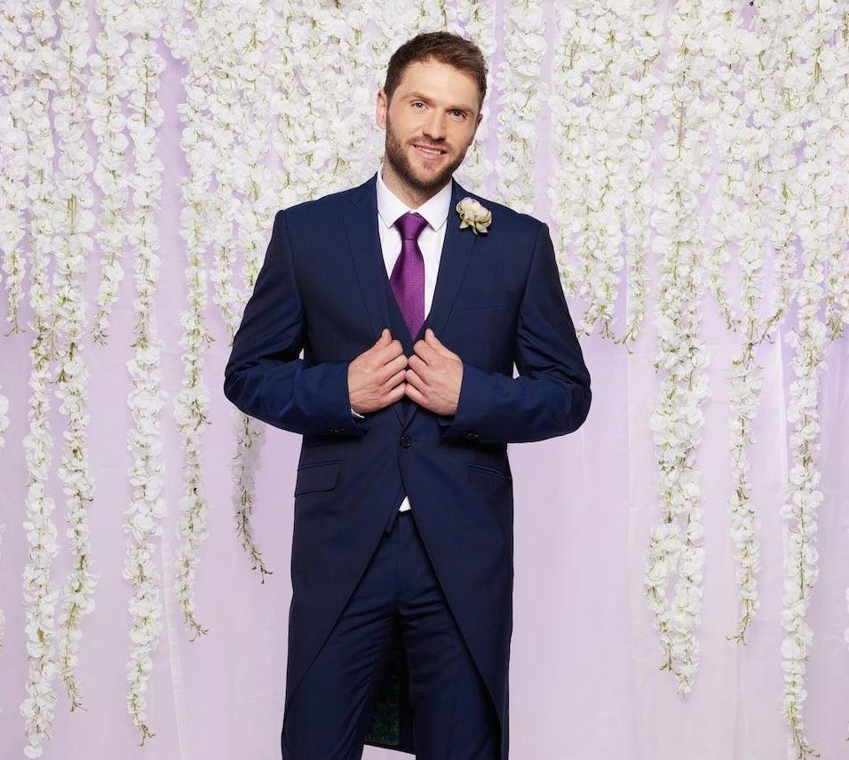 mafs uk 2023 contestant arthur holds the lapels of his navy blue wedding suit and purple tie as he stands and smiles in front of a lilac backdrop with white hanging flowers