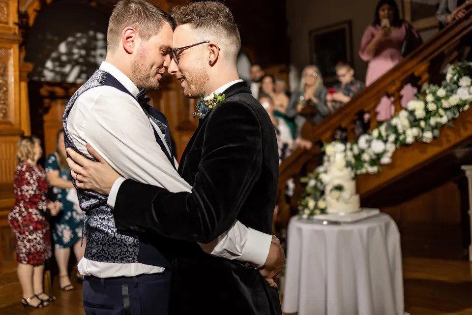 Two grooms hold each other close during their first dance with guests watching in the background