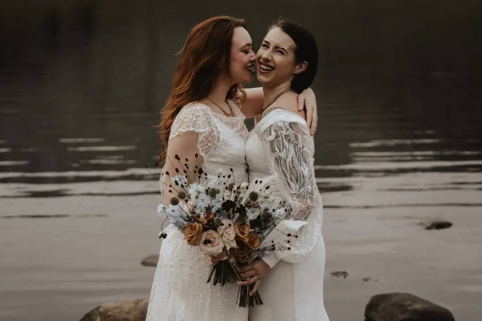 two brides standing in front of a lake laughing at their funny wedding vows