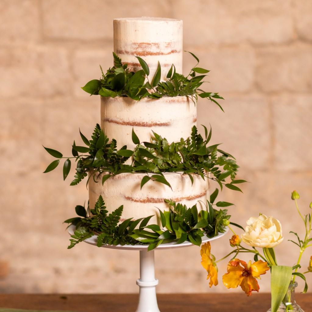 A semi-naked buttercream sponge wedding cake on a cake stand decorated with lots of foliage.