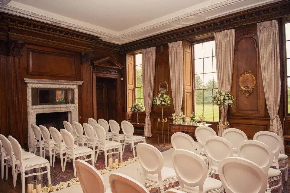 Wood-panelled room set for wedding venue with white chairs, a white aisle lined with flowers and candles, white floral displays, a fireplace and curtains