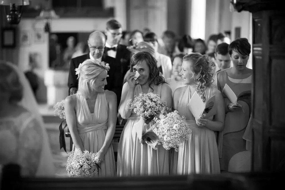 A bridesmaid laughing as she is photographed wiping a tear away during a wedding ceremony, whilst the two other bridesmaids either side look on and giggle