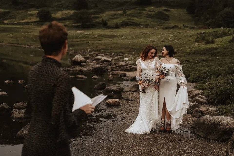 A wedding celebrant awaits two brides with bouquets walking through the countryside