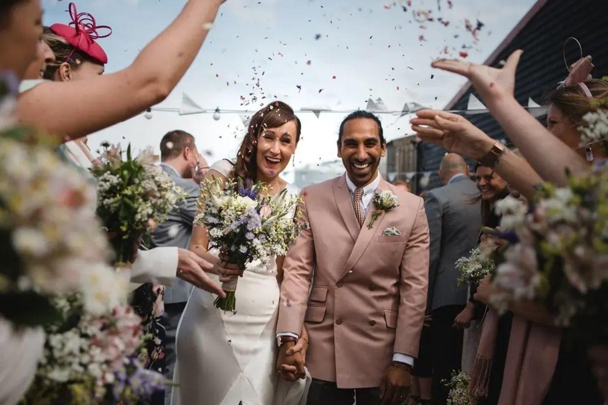 a bride in a wedding dress and a groom in a pink wedding suit walking through the line of recession as guests throw confetti