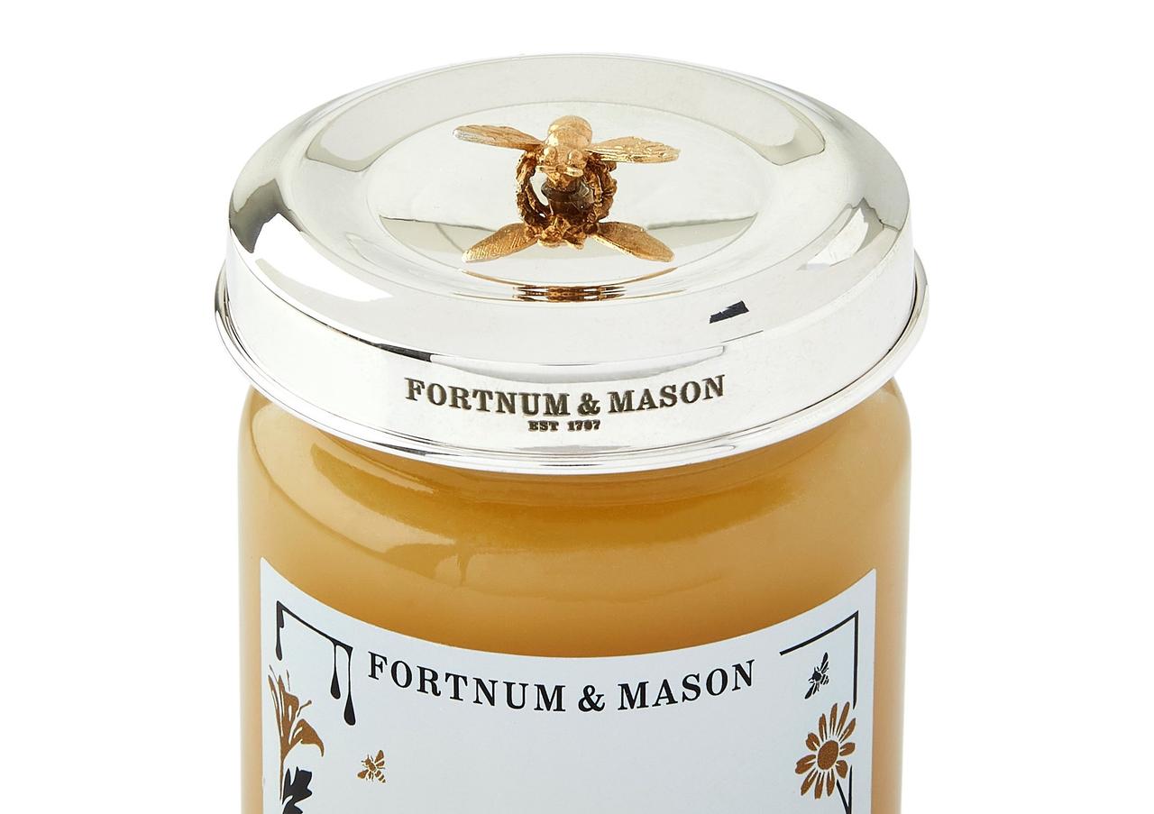 A jar of honey with a sterling silver lid and a decorative gold bee on top