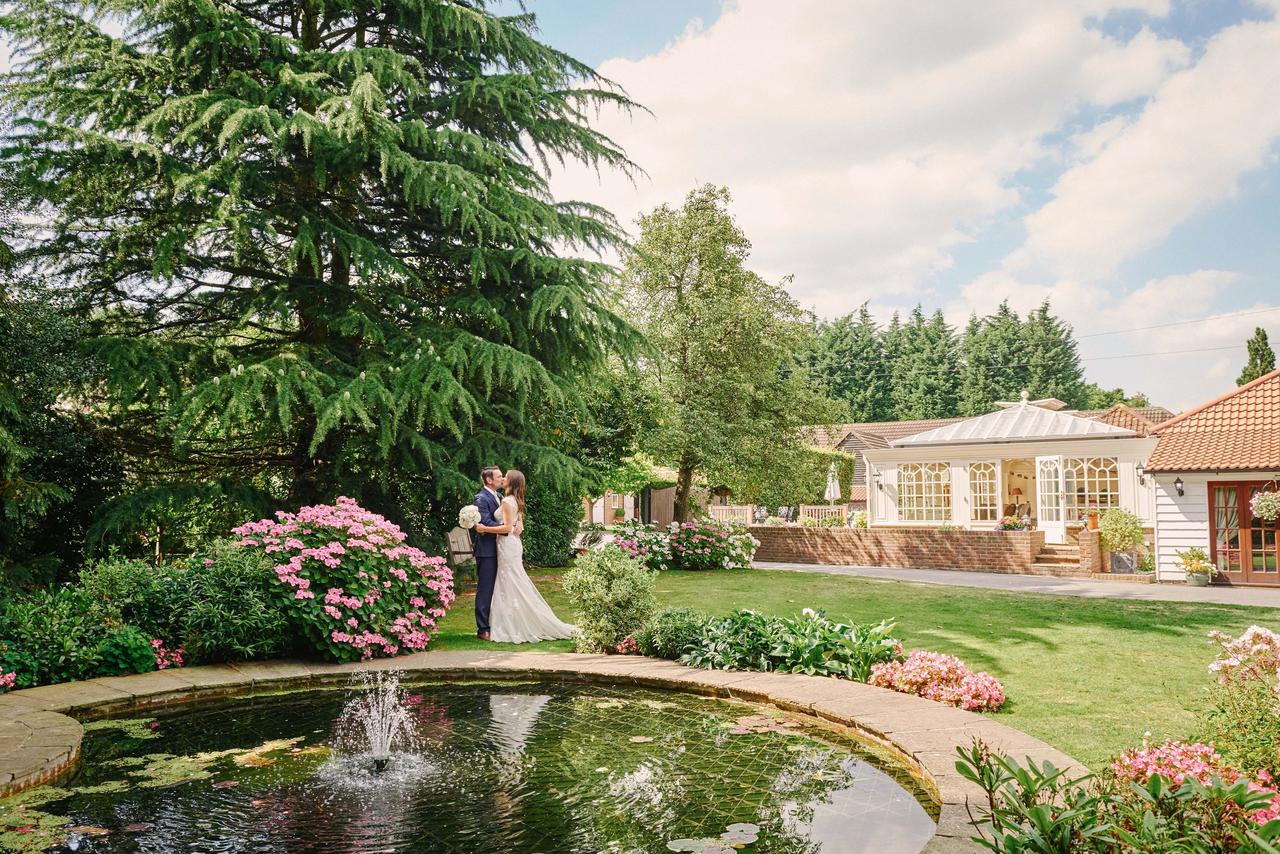 Couple kissing in wedding clothes next to a floral adorned pond at a barn wedding venue