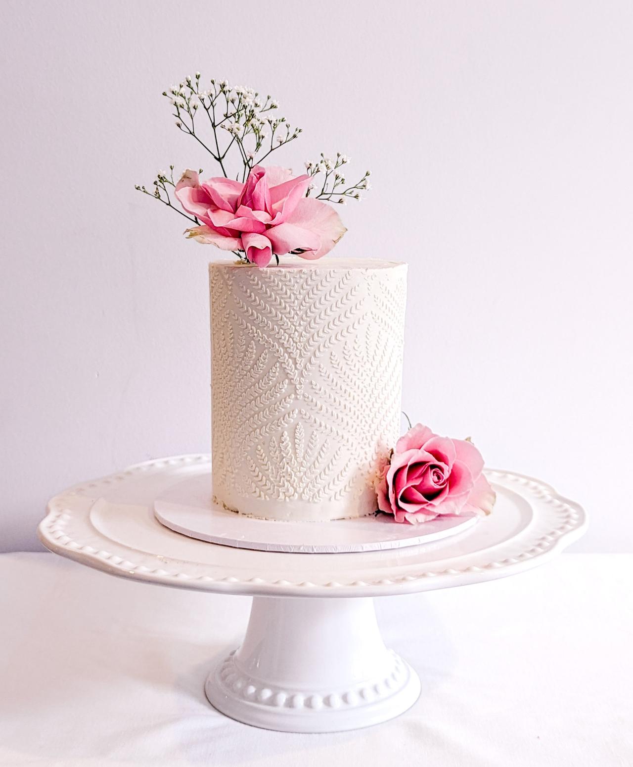 A white single tier wedding cake with lace-textured detail and pink flowers.
