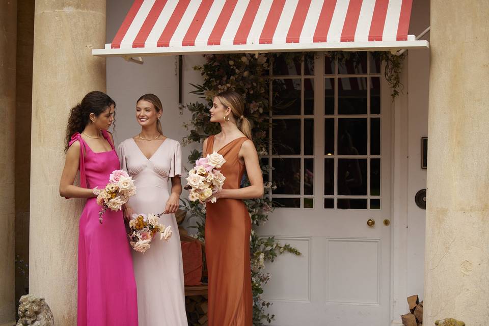 Three girls stood outside a pink door wearing mismatched bridesmaid dresses and holding bouquets, one dress is hot pink with bow tie shoulders, another is short sleeved in pale pink and the third is a cowl-neck gown in burnt oragne