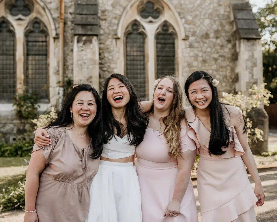 A bride laughing with her head back next to the mother of the bride and two bridesmaids, all with their arms around each other laughing and smiling