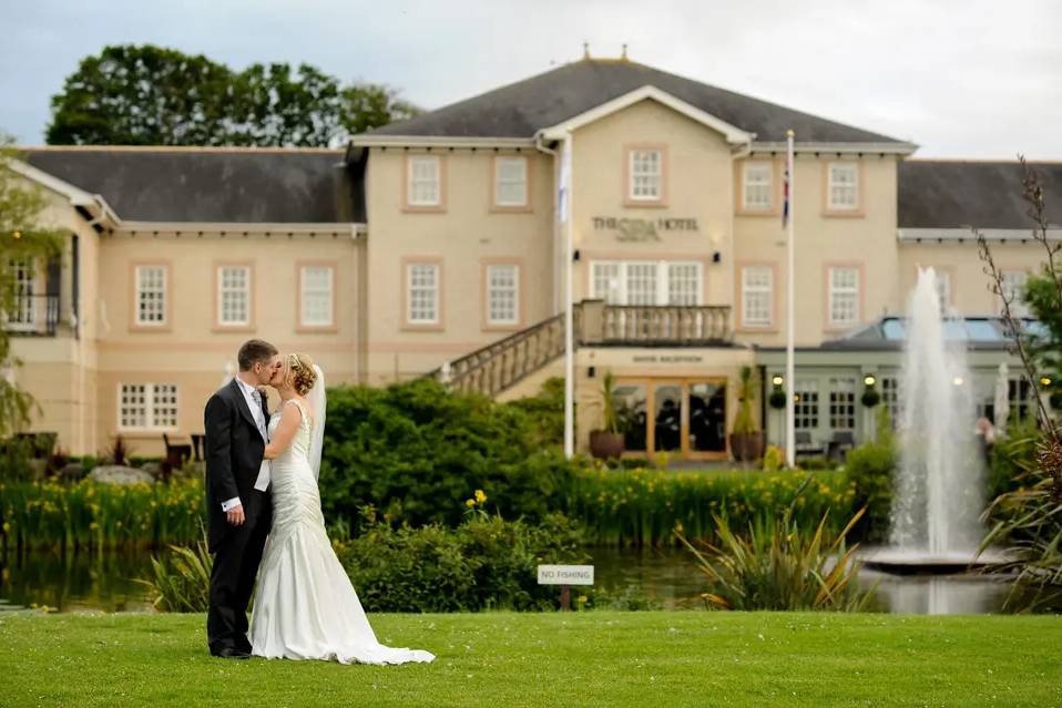 A couple dressed in a suit and wedding dress kisses outside Ribby Hall Village with the fountain going and the plants in bloom