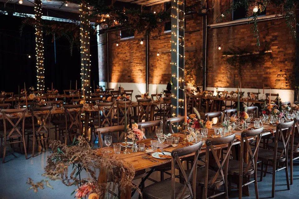 Room set for wedding meal with long wooden tables, wooden chairs, fairy lights wrapped around industrial beams and dried floral arrangements