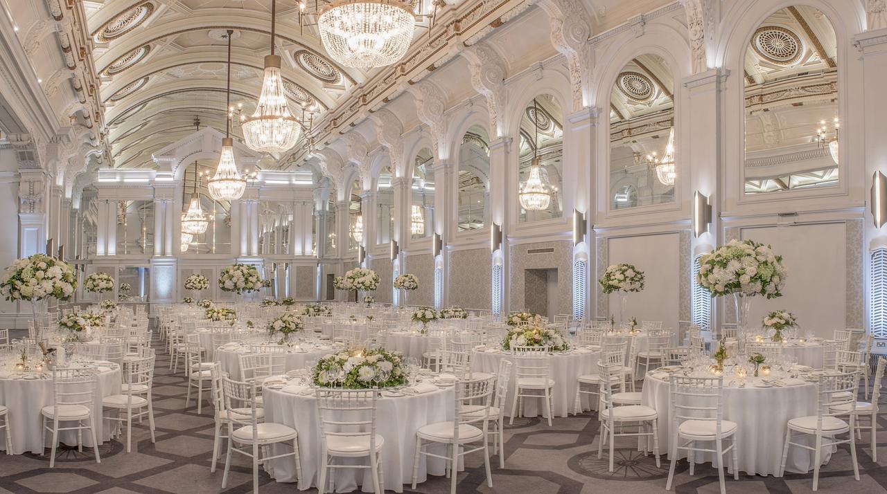 Interior of a ballroom wedding venue set up for a reception with round tables and tall floral centrepieces.