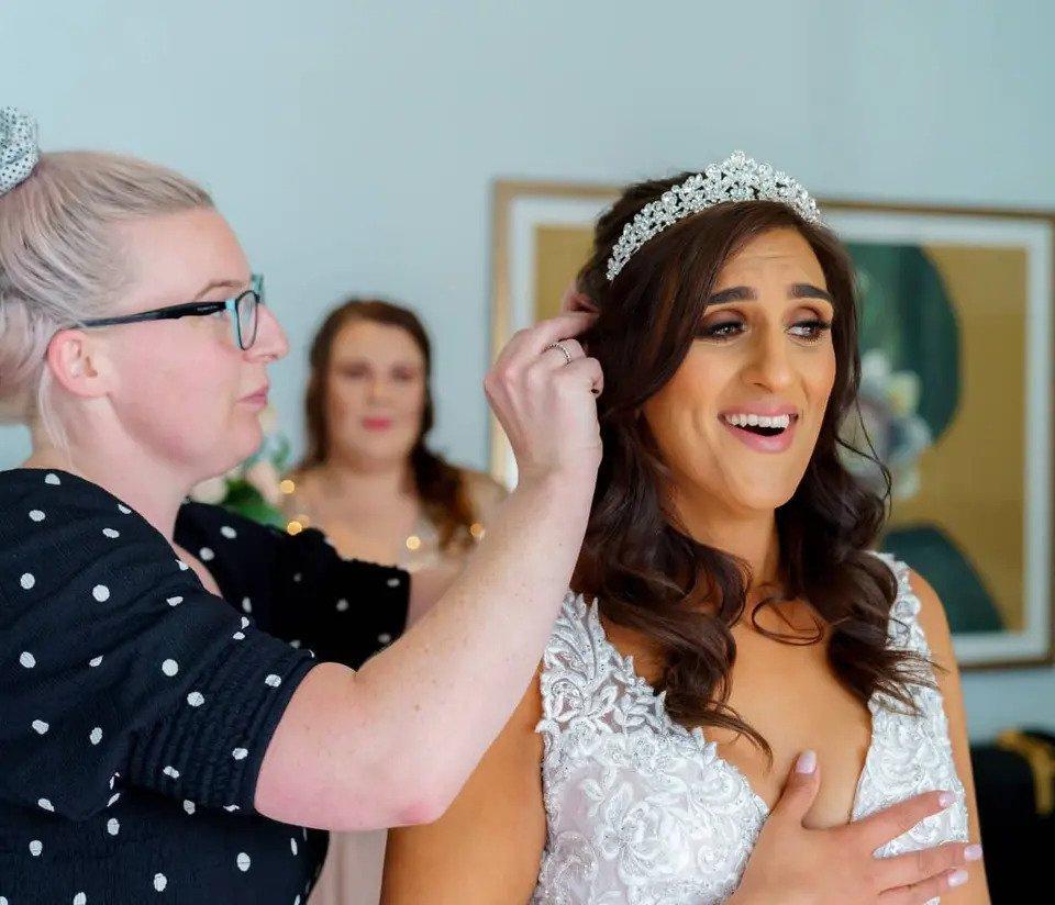 A standing bride with full makeup clasps her hand to her heart while a stylist fixes her hair