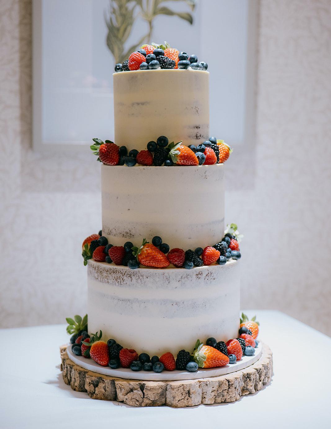 A three tiered white semi naked cake decorated with fresh fruit on a log slice serving board.