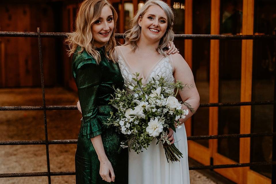 A bride wearing a beaded v-neck wedding dress standing with her bridesmaid who is dressed in a shiny green bridesmaid dress and gold block heels outside the wedding venue