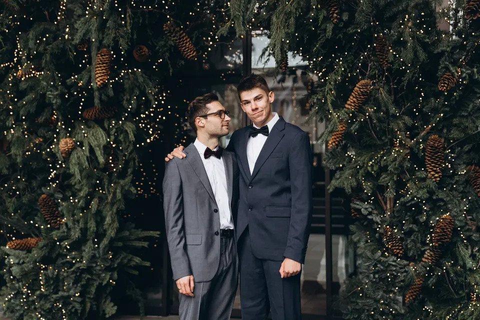 Two grooms stand under foliage and fairylights