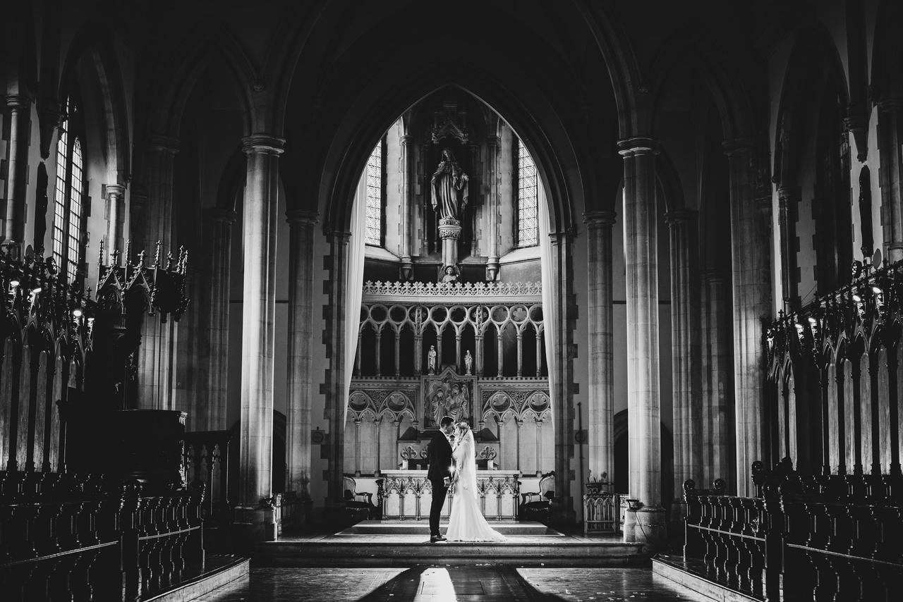 A black and white photograph of the inside of a high-ceilinged chapel with gorgeous architectural detail and a couple standing at the altar saying their vows